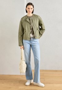 Giacca bomber verde oliva con cintura in vita, abbinata a jeans a zampa d'elefante azzurri chiari e sneakers bianche. Tiene una borsa a tracolla color crema.