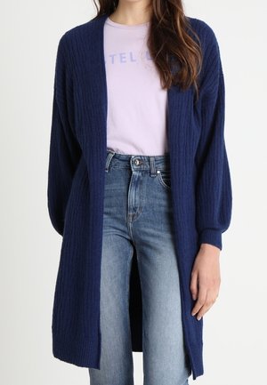 Vrouw die een lange marineblauwe geribbelde cardigan draagt over een lichtpaarse grafische t-shirt en hooggetailleerde vervaagde blauwe spijkerbroek.