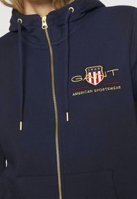 Námořnická modrá mikina na zip vyrobená z bavlny, s přírodními zlatými zipy, vyšitým logem na hrudi a stahovací kapucí.