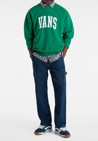 Felpa verde con logo "VANS", indossata sopra una camicia a scacchi grigia e nera, abbinata a jeans blu larghi e sneakers multicolori.