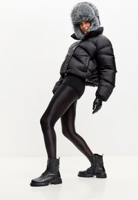 Chaqueta de plumas negra con capucha de piel gris, combinada con leggins negros brillantes y botas de combate negras con cordones. Guantes de cuero.