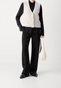 Person, der ein schwarzes Shirt, schwarze weite Hosen, eine cremefarbene, flauschige Weste trägt, schwarze spitze Schuhe anhat und in einer Hand eine cremefarbene Handtasche hält.
