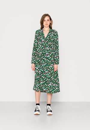 Vestido com padrão floral, base verde, mangas compridas e gola. Usado com sapatilhas pretas e brancas. A bainha cai abaixo do joelho.