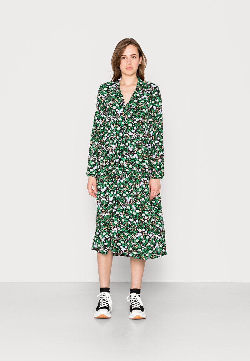 Vestido com padrão floral, base verde, mangas compridas e gola. Usado com sapatilhas pretas e brancas. A bainha cai abaixo do joelho.