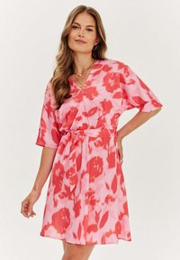 Kratký rukáv šaty v růžové barvě s červenými květinovými vzory, s překládaným designem a opaskem na uvázání v pase. Hladká, lehká látka.