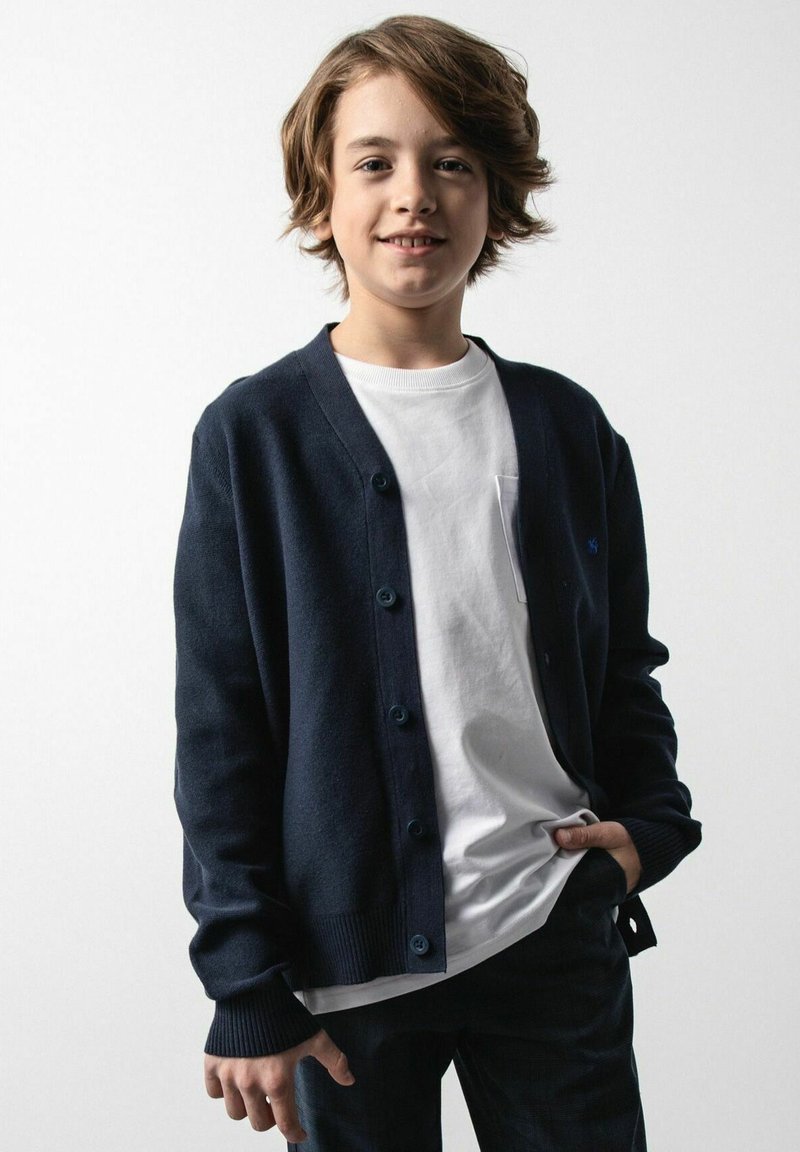 Granatowy kardigan z okrągłym dekoltem, zapinany na guziki, z prążkowanymi mankietami, noszony na białej koszuli. Koszula z kieszonką, luźny krój.