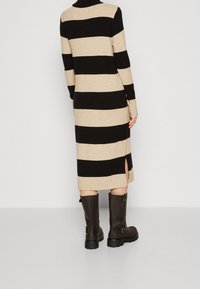 Stickad klänning med vertikala svarta och beige ränder, långa ärmar, hög halsringning och sidslitsar; kombinerad med knähöga bruna läderstövlar.