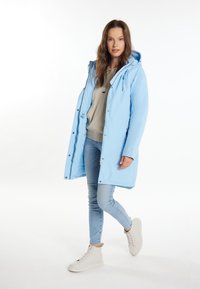 Parka impermeabile azzurra chiaro con cappuccio, dotata di bottoni automatici, tasche laterali e vestibilità comoda, abbinata a jeans di denim chiaro e sneakers beige.