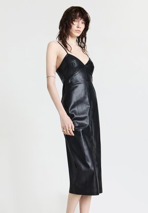Donna con i capelli mossi e scuri, che indossa un vestito midi in pelle nera aderente con spalline sottili, in piedi su uno sfondo bianco uniforme.