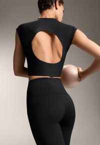 Črni športni komplet vključuje oprijet vrh z velikim ovalnim izrezom na hrbtu in hlačne nogavice z visokim pasom, narejene iz gladkega, raztegljivega materiala.