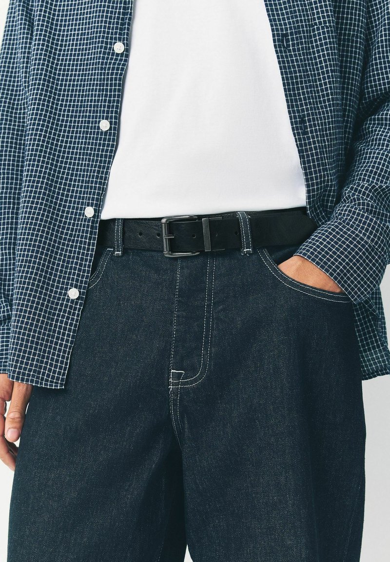 Cinturón de cuero negro con una hebilla metálica rectangular, combinado con una camisa de cuadros en tonos azul marino y pantalones de mezclilla oscuros con detalles en costura blanca.