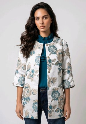 Mujer con cabello largo y oscuro que lleva una chaqueta blanca con flores con rosas azules y beige sobre una blusa verde azulado con volantes y vaqueros oscuros.