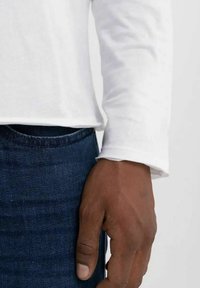 Camicia bianca in cotone a maniche lunghe con colletto tondo, abbinata a jeans in denim blu scuro. Mano visibile appoggiata sui fianchi.