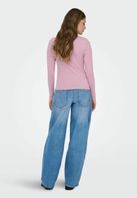 Frau mit langen, welligen Haaren, die ein rosa Langarmoberteil und lockere, hellblaue Jeans trägt, steht und ist mit dem Rücken zu sehen vor einem weißen Hintergrund.