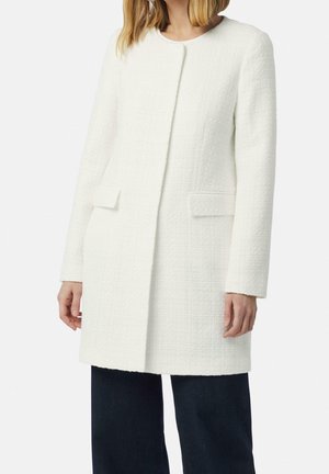 Woman wearing a white textured long coat with flap pockets over dark wide-leg pants, standing against a plain background.