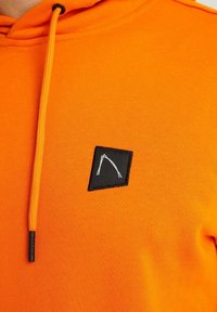 Felpa arancione brillante con un patch quadrato nero che presenta un design geometrico minimalista bianco sul petto e un cordino arancione abbinato.