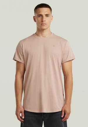 Uomo con capelli corti che indossa una maglietta rosa chiaro con le maniche arrotolate e jeans scuri, in piedi davanti a uno sfondo beige uniforme.