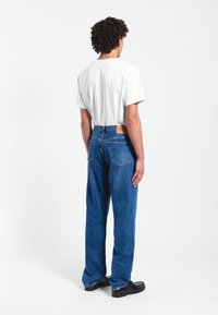 Vaqueros de mezclilla azul con un corte relajado, que presentan un parche de cuero marrón en la cinturilla trasera. Combinados con una camiseta blanca lisa.
