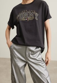 Černé tričko s krátkým rukávem s nápisem "TOMMY New York" tištěným zlatou a bílou barvou, doplněné lesklými stříbrnými kalhotami s širokými nohavicemi. Viditelná texturová látka.
