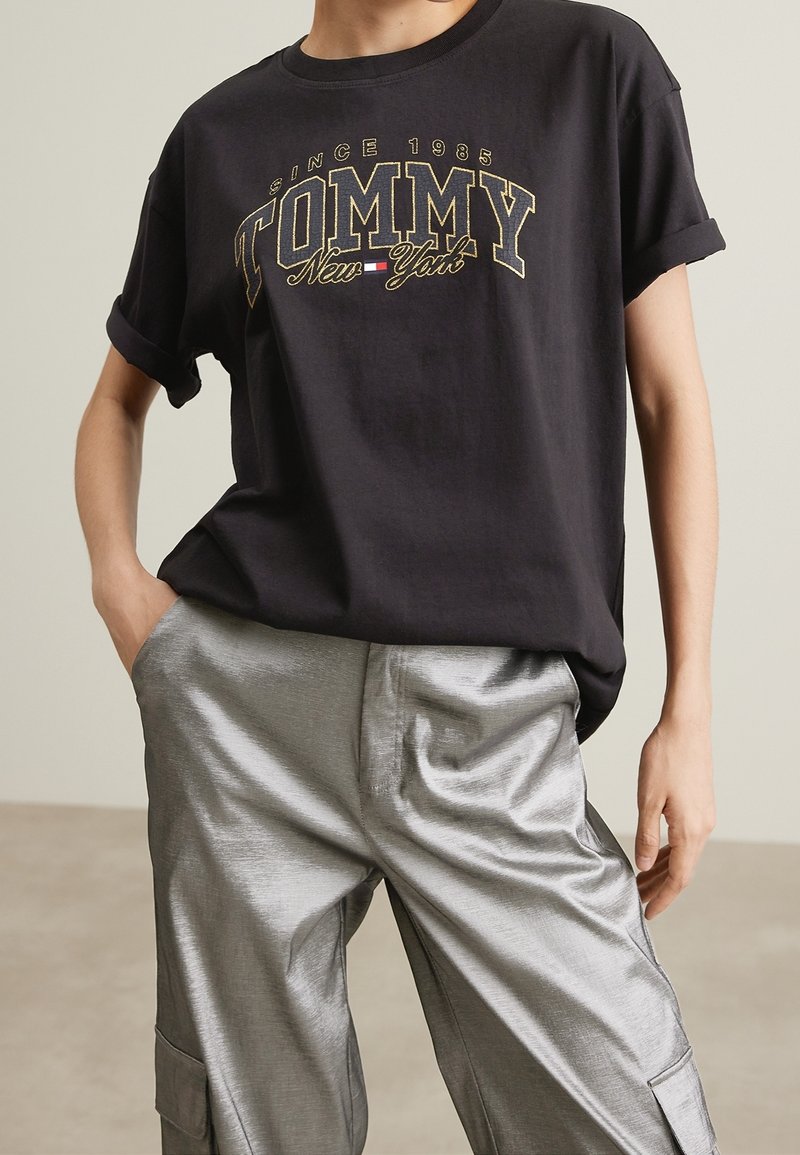 Černé tričko s krátkým rukávem s nápisem "TOMMY New York" tištěným zlatou a bílou barvou, doplněné lesklými stříbrnými kalhotami s širokými nohavicemi. Viditelná texturová látka.