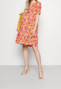 Florales Kleid mit lebhaften Mustern in Orange, Pink und Gelb. Kurze Ärmel, V-Ausschnitt und ein ausgestellter Rock. Getragen mit beigen Sandalen mit dicken Absätzen.