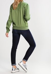 Veste bomber verte avec poignets et ourlet côtelés, associée à un jean skinny bleu foncé et des baskets argentées métalliques. Texture lisse sur toute la surface.