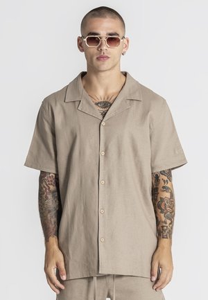 Muž s krátkymi vlasmi a slnečnými okuliarmi, oblečený v béžovej krátkorukávovej košeli s gombíkmi, ukazujúci tetovania na hrudi a oboch rukách na jednoduchom pozadí.