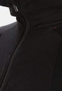 Giacca in pile nera con cerniera e collo alto, caratterizzata da una texture liscia, cuciture minimali e una piccola etichetta del marchio vicino al cappuccio.