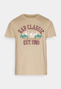 Beżowa bawełniana koszulka z okrągłym dekoltem, z grafiką gór i drzew, z napisem "GAP CLASSIC" oraz "EST. 1969" w bordowym kolorze.