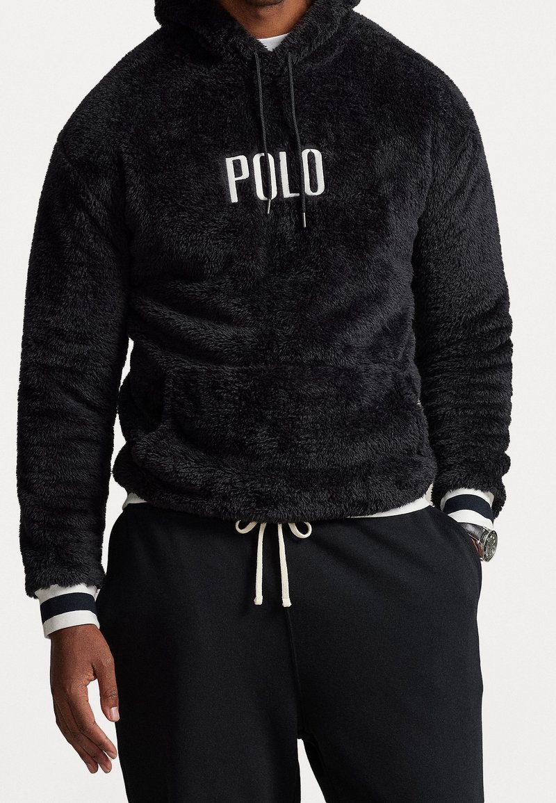 Svart plyschhoodie med "POLO"-logotyp, med känguruficka, snörning i huvan och randiga muddar över en vit skjorta.