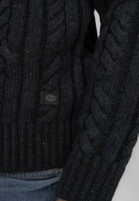 Dunkelblauer Kabelstrickpullover mit dicken, strukturierten Mustern. Gerippte Bündchen und Saum. Kleines schwarzes Lederetikett mit dem Markennamen auf der Vorderseite.
