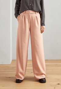 Světle růžové kalhoty s širokými nohavicemi a sklady v pase, zkombinované s černo-bílou pruhovanou košilí a tmavohnědými botami. Hladká textura.