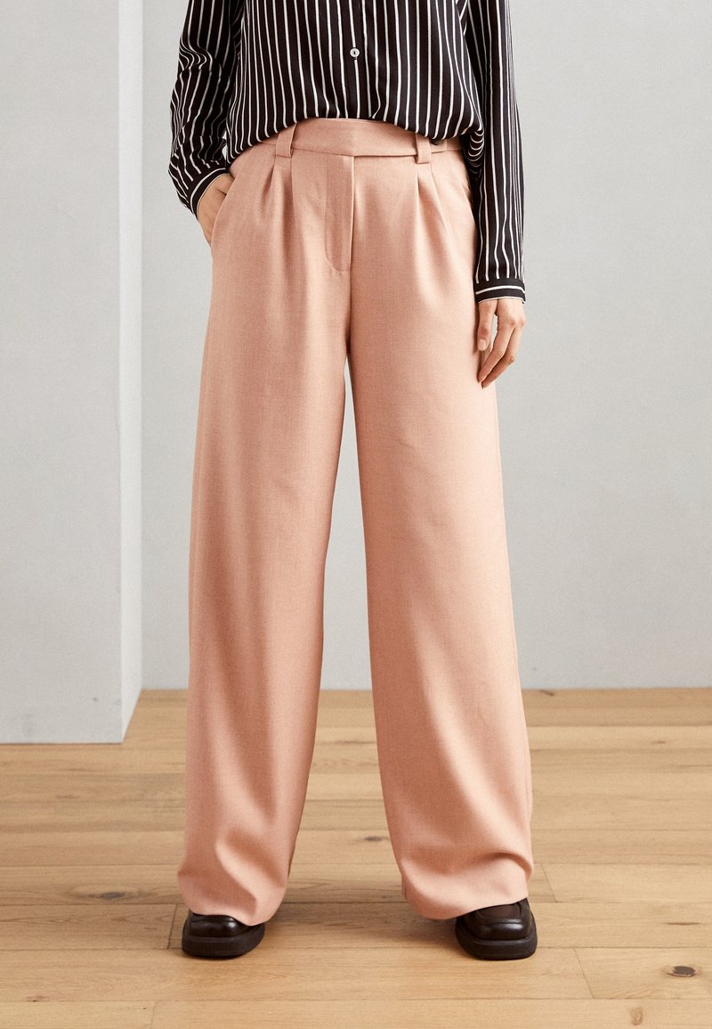 Světle růžové kalhoty s širokými nohavicemi a sklady v pase, zkombinované s černo-bílou pruhovanou košilí a tmavohnědými botami. Hladká textura.