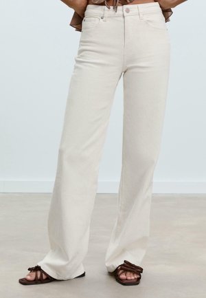 Femme portant un jean écru taille haute à jambes larges et des sandales marron à brides, debout sur un sol gris devant un fond uni.