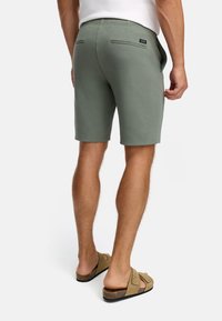 Mand iført olivenfarvede shorts og brune slip-on sandaler, står med ryggen til på en ensfarvet hvid baggrund.