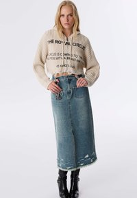 Pull à capuche cropped beige avec texture côtelée et texte, associé à une longue jupe en denim bleu clair, avec un ourlet effiloché et une poche avant.