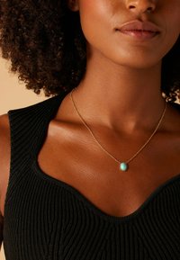 Gouden ketting met een ovale turquoise hanger, gedragen met een zwarte geribbeld top met een wijd decolleté, benadrukkend de textuur en kleurcontrast.