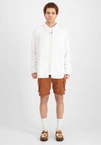 Chemise blanche à manches longues avec boutons, short cargo marron, chaussettes blanches, chaussures marron à motifs, se tenant contre un fond uni.