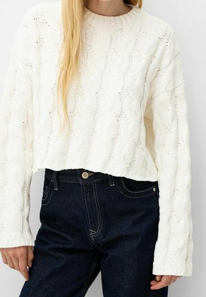 Kvinde iført en hvid kabelstrikket cropped sweater og mørkeblå højtaljede jeans, vist fra skuldre til hofter.