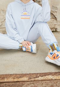 Felpa leggera azzurra con un logo arcobaleno, abbinata a pantaloni della tuta coordinati e scarpe bianche con accenti arancioni, blu e grigi.