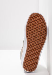 Semelle d'une basket beige et blanche montrant une semelle extérieure en caoutchouc marron à motif gaufré sur un fond blanc uni.
