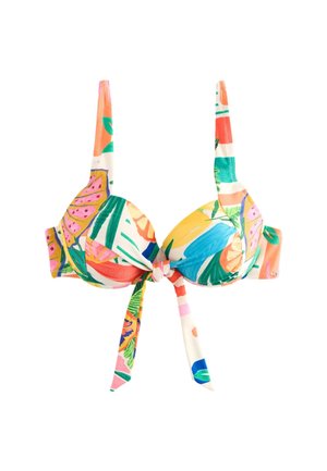 Femme en bikini coloré à motif de fruits et short, debout pieds nus sur une plage de sable, tenant une chaise en plastique orange près de l'océan.