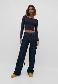 Un haut crop à manches longues bleu marine associé à un jean cargo bleu foncé avec deux poches latérales et des détails en zip. Le modèle porte des chaussures jaunes.