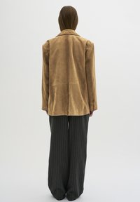 Bruine suede blazer met een revers en een verlengd achterpand, gecombineerd met een donkergrijze broek met brede strepen. Soepele textuur.
