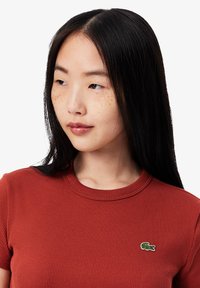 T-shirt rouge à rayures, à manches courtes, avec un col rond et un petit logo vert Lacoste sur la poitrine gauche. Texture lisse.