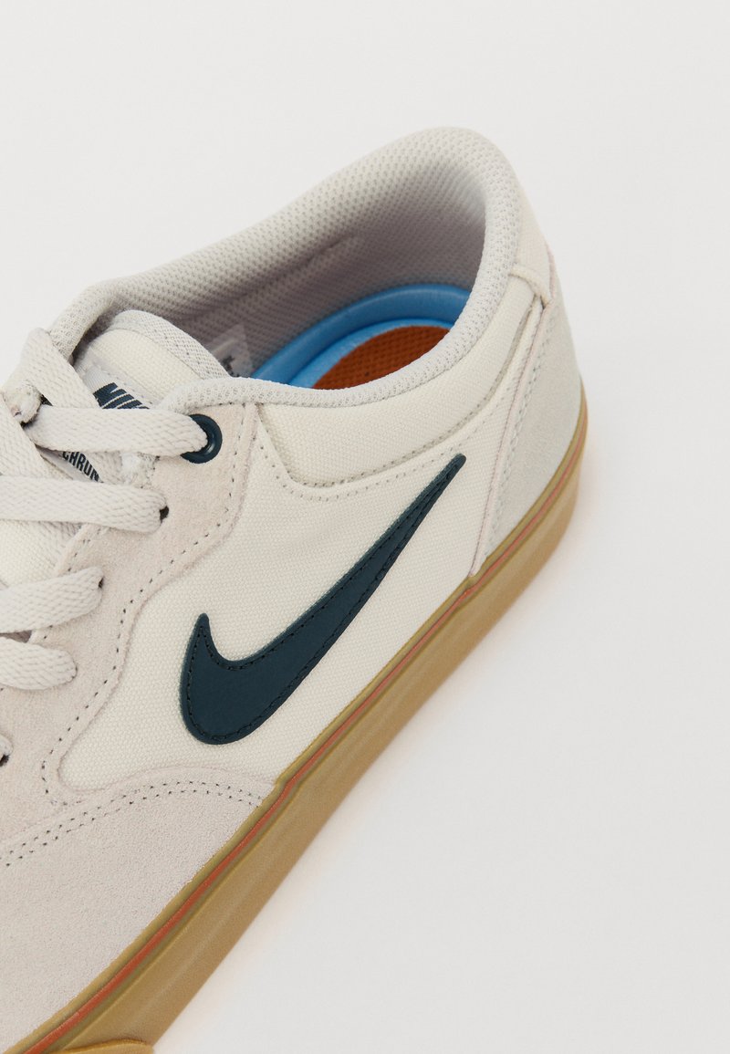 Zapatilla con parte superior de lona beige, plantilla azul y logo de Nike en azul oscuro. Cuenta con una suela de goma y detalles texturizados en la parte superior.