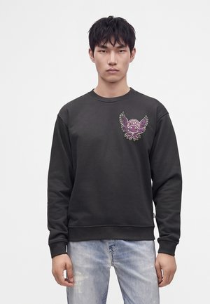 Jeune homme aux cheveux noirs portant un sweat-shirt noir avec un emblème ailé violet et un jean bleu clair sur un fond uni.