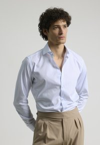 Camisa blanca con rayas verticales azul claro, cuello abotonado y mangas largas, combinada con pantalones beige. Textura suave.