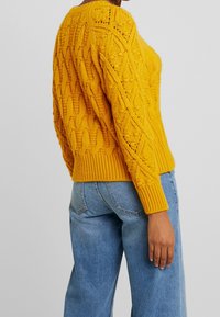 Gul stickad tröja med texturerade kabelmönster, ribbad nederkant och ärmslut, parat med ljusblå jeans. Tydliga sömnadsdetaljer synliga.