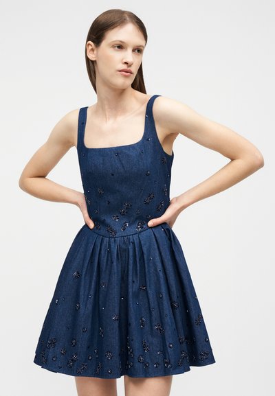 Νέα γυναίκα που φοράει αμάνικο μπλε φόρεμα με πούλιες σε σχέδια λουλουδιών, στέκεται με τα χέρια στη μέση μπροστά σε ένα απλό φόντο.